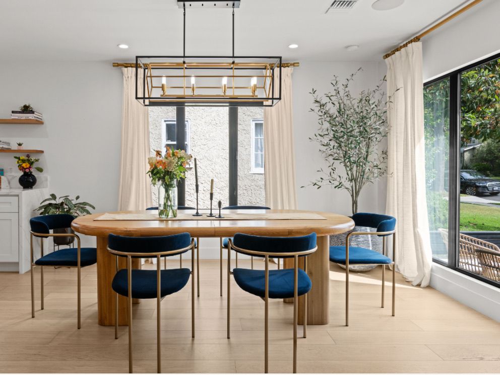 Dining room with a rounded white oak table, navy velvet chairs with brass frames, a black and brass linear chandelier, linen drapery and a potted olive tree