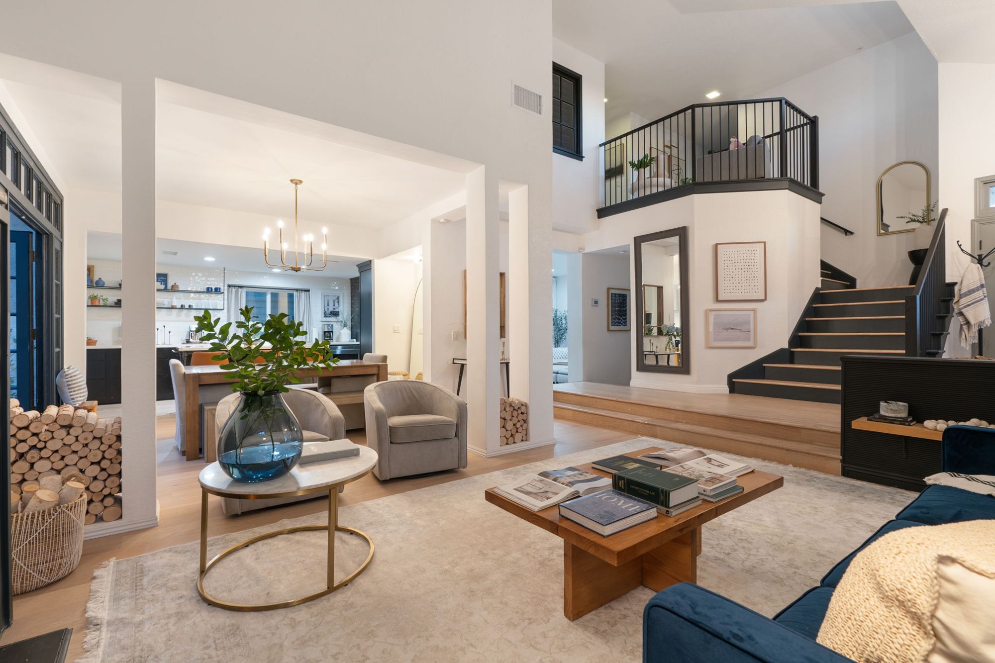 Double-height great room with a navy velvet sofa, wood coffee table stacked with books, brass side table, stacked birch logs and a black stair with oak treads