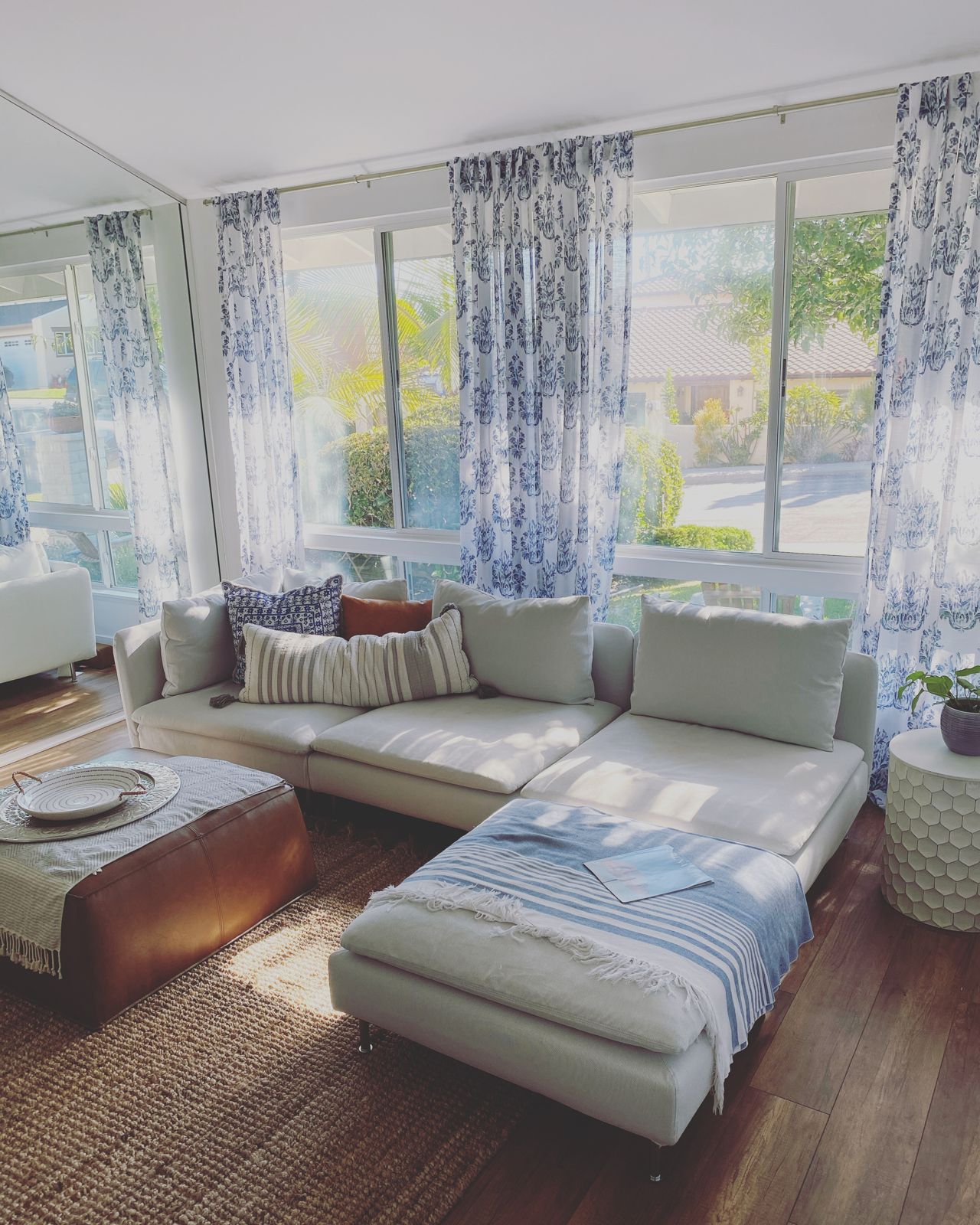 Sunlit living room with a linen sectional, blue block-print curtains, a leather ottoman and jute rug